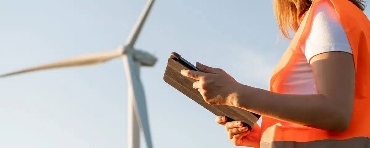 Female an engineer in a helmet and an orange vest controls the operation of a wind turbine using a tablet on the sky background