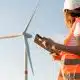 Female an engineer in a helmet and an orange vest controls the operation of a wind turbine using a tablet on the sky background